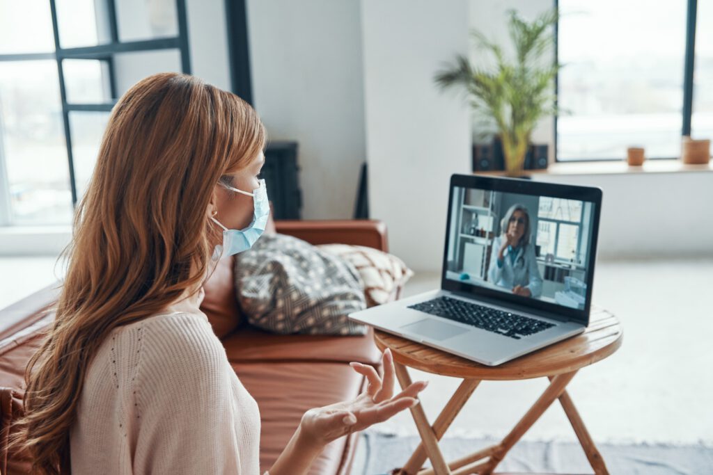 Young woman in protective face mask talking to doctor while having a video conference on laptop